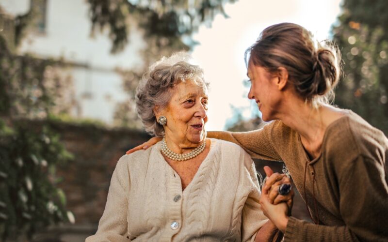 elderly woman with short gray hair wearing a beige sweater talking to a female caregiver wearing a brown sweater