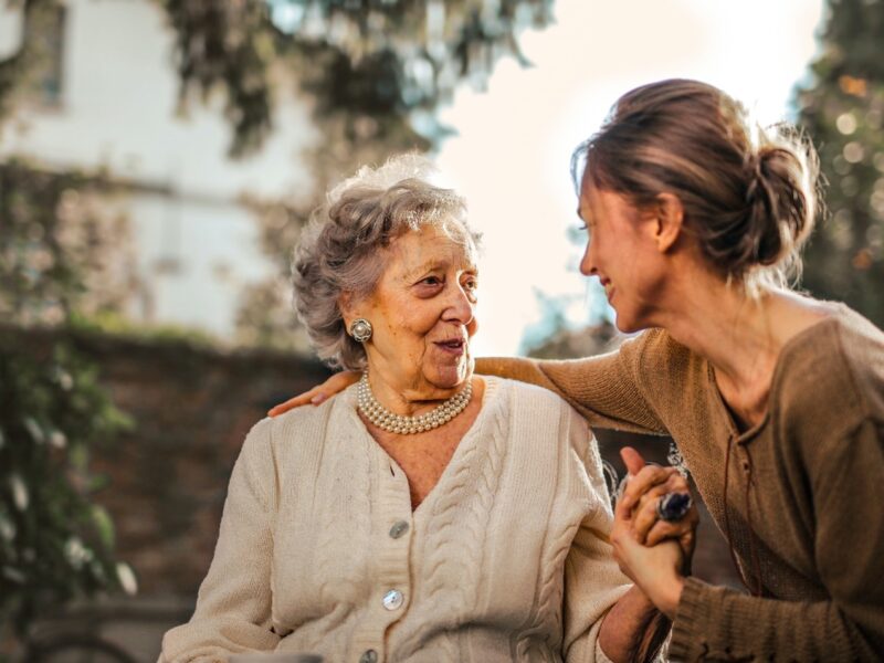 elderly woman with short gray hair wearing a beige sweater talking to a female caregiver wearing a brown sweater