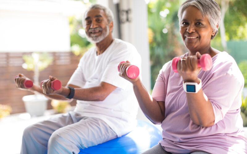 senior woman in a pink tee-shirt and a senior man in a white tee-shirt lifting pink dumbbells while doing seated exercises