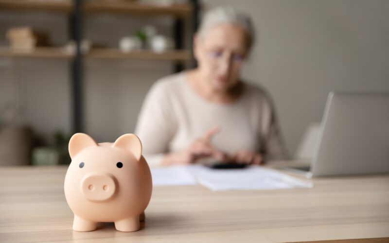 pink piggy bank on a wood-topped desk with a white-haired senior in a tan top working on a calculator in the background