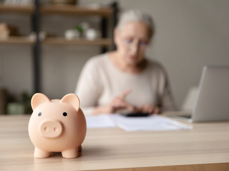 pink piggy bank on a wood-topped desk with a white-haired senior in a tan top working on a calculator in the background