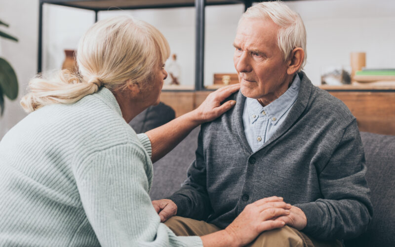 older blond woman with a ponytail and light bluish-gray sweater holds the hand of a white-haired man in a gray sweater