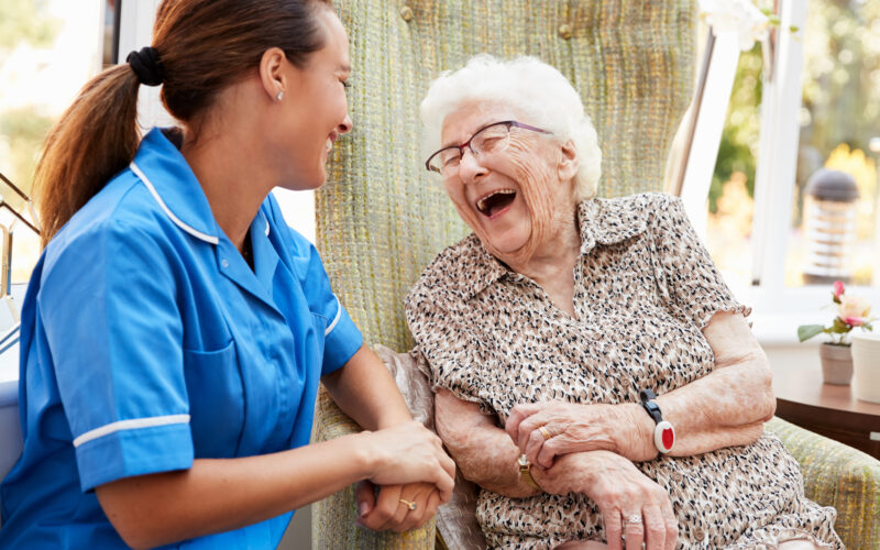Senior Woman Sitting In Chair And Talking With Nurse In Retirement Home