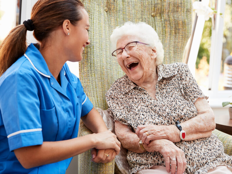 Senior Woman Sitting In Chair And Talking With Nurse In Retirement Home