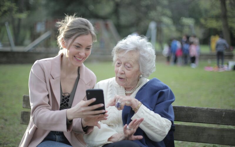 smiling white haired woman wearing an ivory sweater and a blue shawl looking at the smartphone shown to her by a smiling blond woman in a pink blazer; both are sitting on a bench with a park-like setting in the background