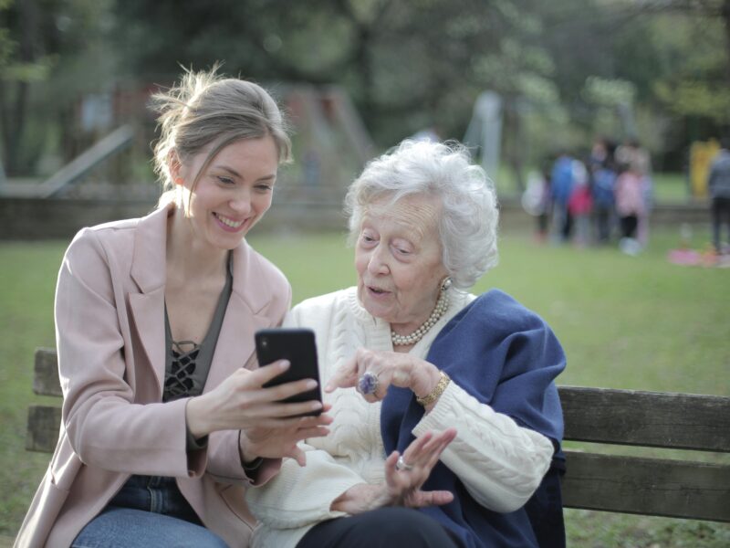 smiling white haired woman wearing an ivory sweater and a blue shawl looking at the smartphone shown to her by a smiling blond woman in a pink blazer; both are sitting on a bench with a park-like setting in the background