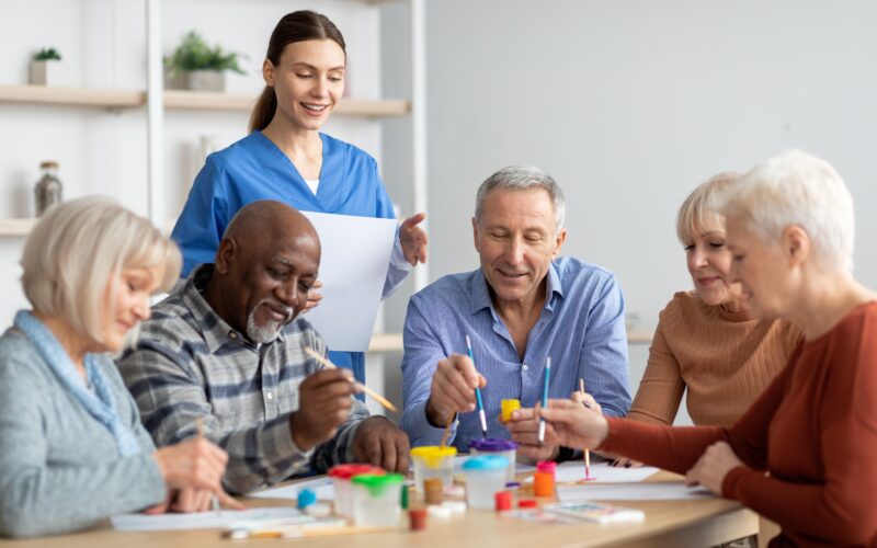 five smiling older adults enjoying the benefits of art for seniors painting at a light wood table in a room with white walls while a woman in a blue scrub top looks on while also smiling