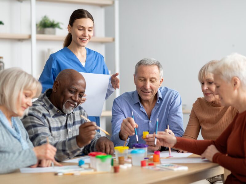five smiling older adults enjoying the benefits of art for seniors painting at a light wood table in a room with white walls while a woman in a blue scrub top looks on while also smiling
